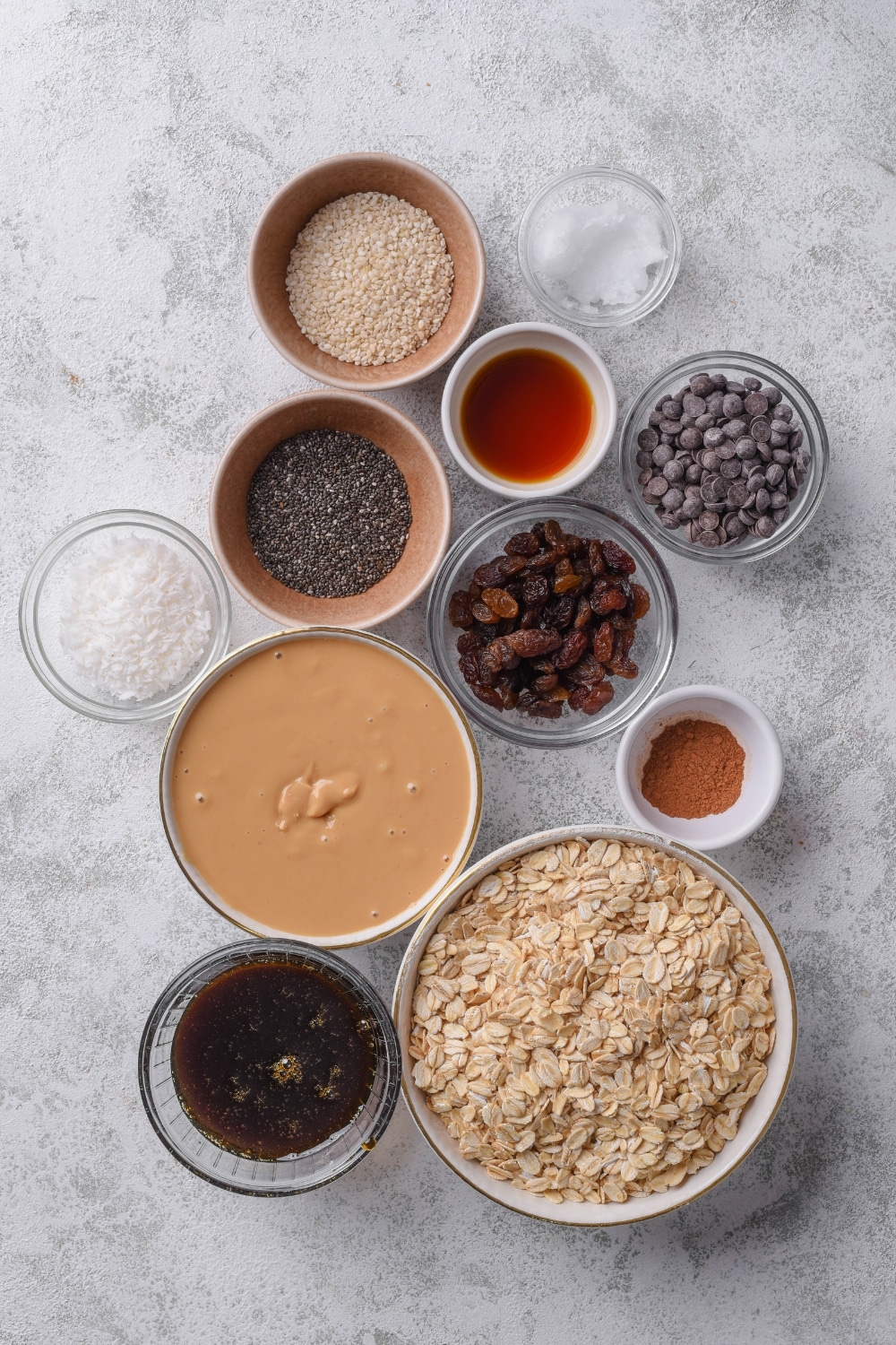 An overhead shot of several bowls containing various low calorie energy bar ingredients like oats, chia seeds, raisins, chocolate chips, peanut butter, shredded coconut, maple syrup, and sesame seeds