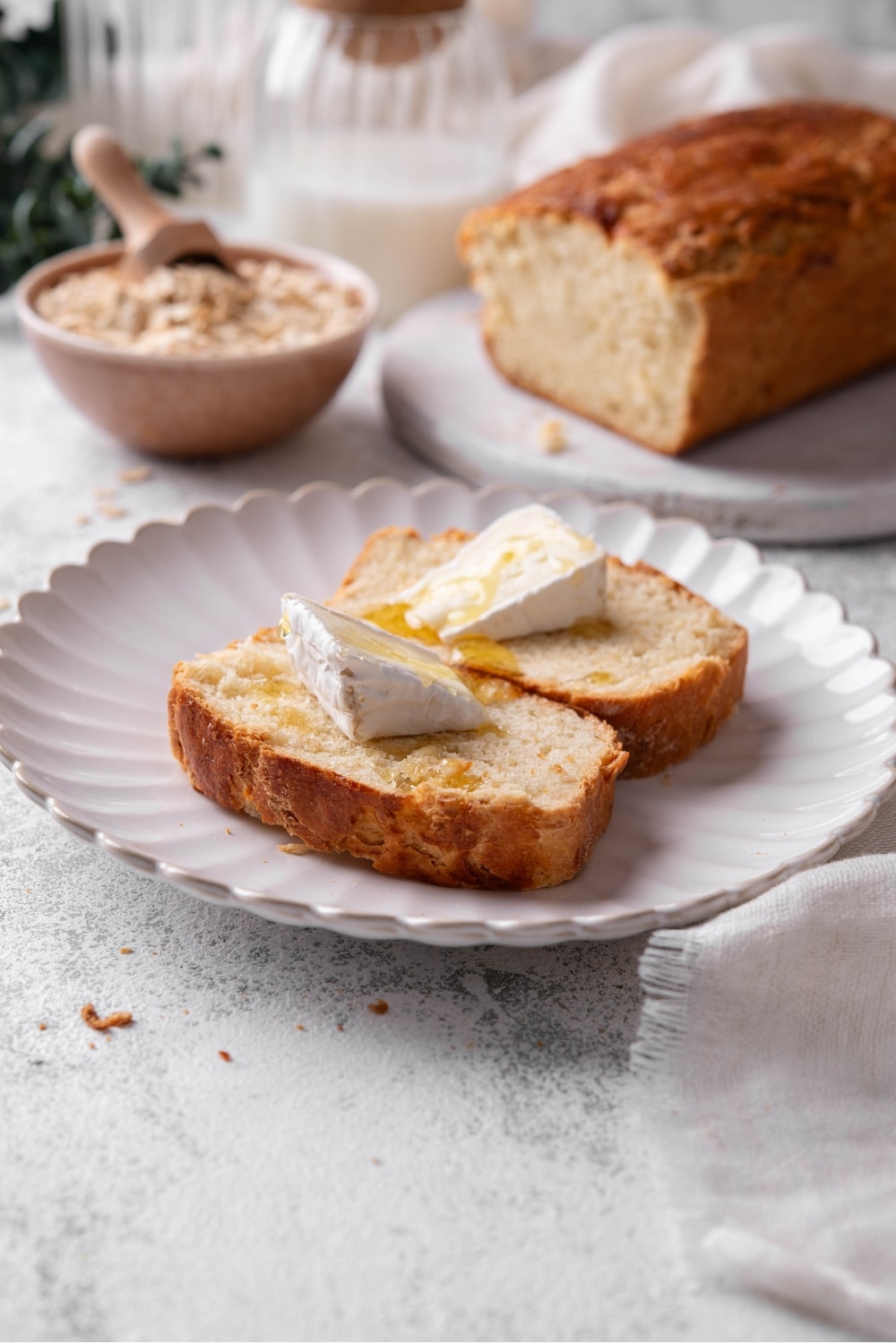 Two slices of oat bread topped with butter and drizzled with honey are on a white ceramic plate. There is an extra loaf of oat bread in the background along with a bowl of oats with a scooper inside it.