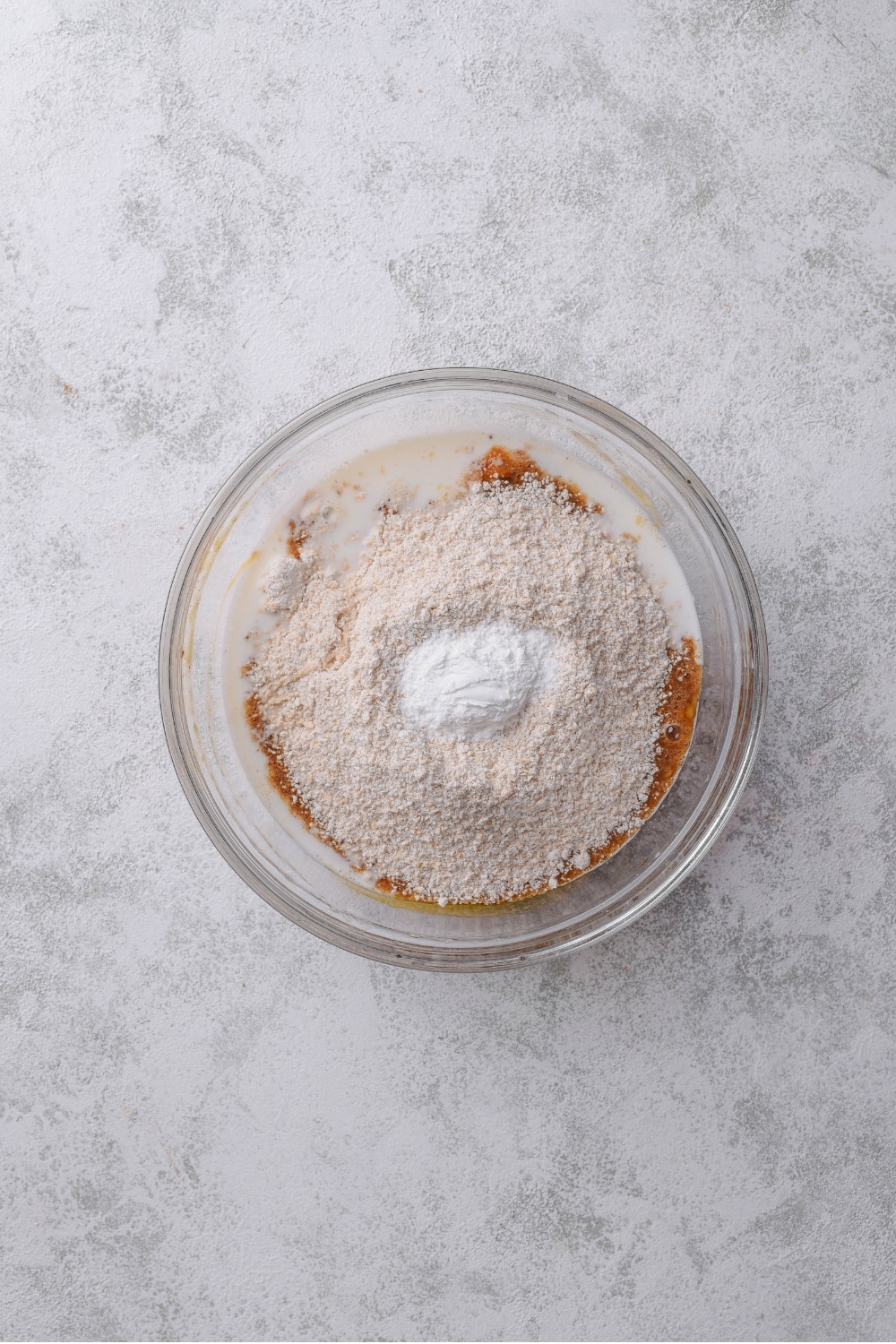 An overhead shot of a glass bowl with a mashed banana mixture topped with flour and baking powder.