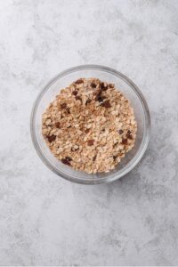 An overhead shot of a glass bowl with oats combined with cinnamon, raisins, chia seeds, and other ingredients on a marble counter.