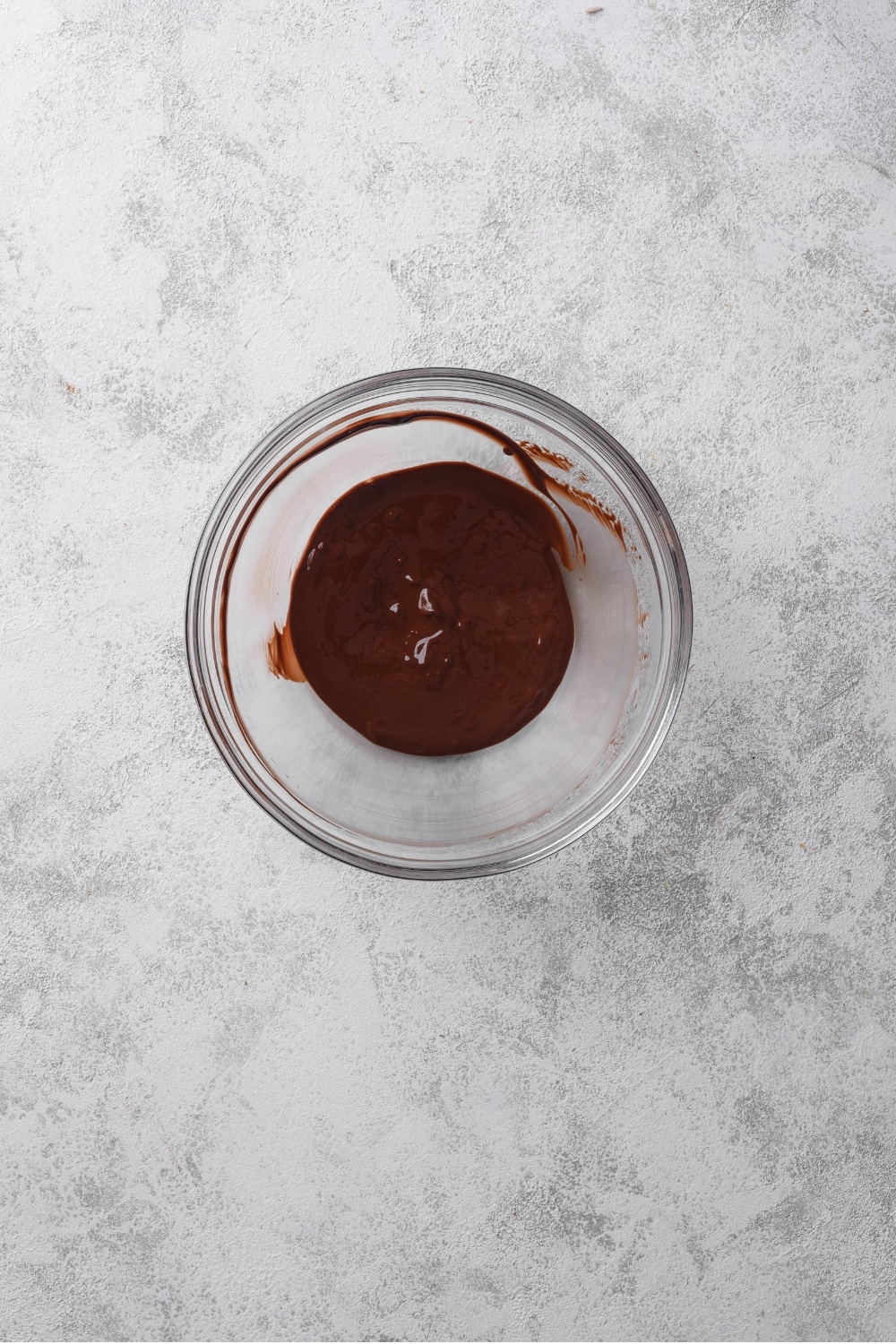 An overhead shot of a glass bowl with melted dark chocolate on a marble counter.