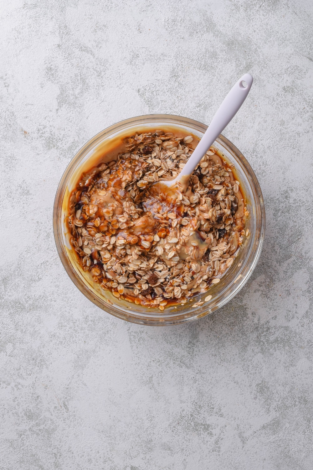 An overhead shot of a glass bowl with an oat mixture combined with peanut butter and syrup. There is a spoon in the bowl stirring the oat mixture