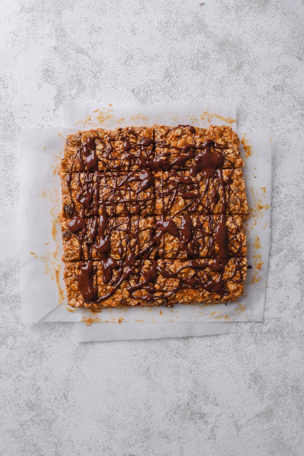 An overhead shot of a batch of uncut energy bars on top of parchment paper with chocolate sauce drizzled on top