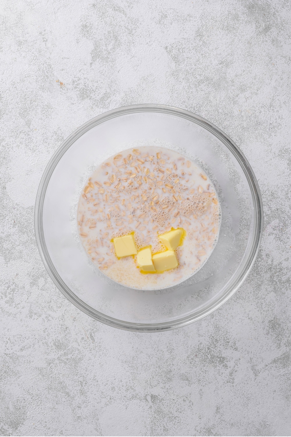 An overhead shot of a glass bowl filled with oats soaking in milk with tabs of butter.