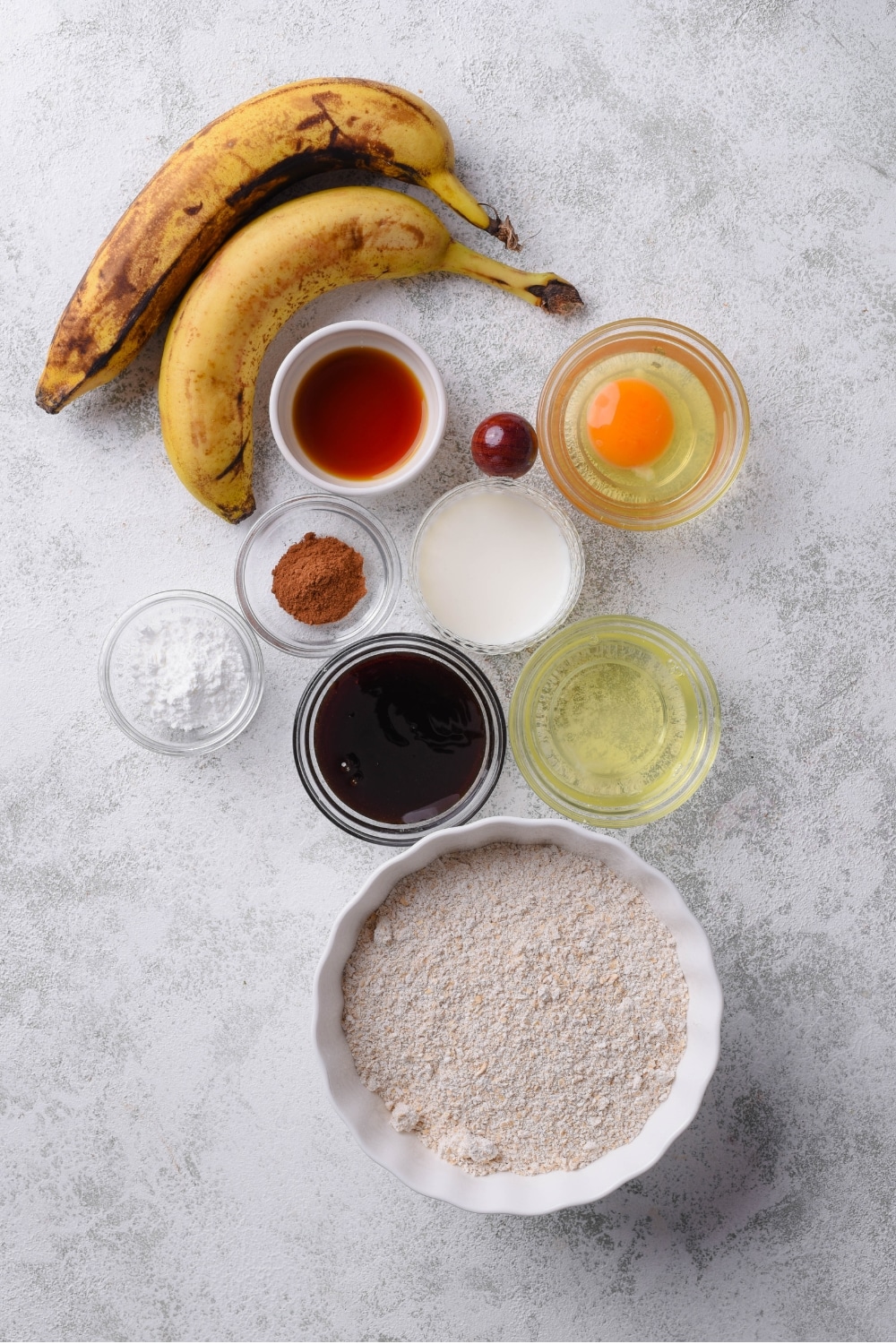 An overhead shot of several bowls of various shapes sizes filled with ingredients to make low calorie banana bread such as oat flour, egg, egg white, vanilla, syrup, cinnamon, and two ripe bananas