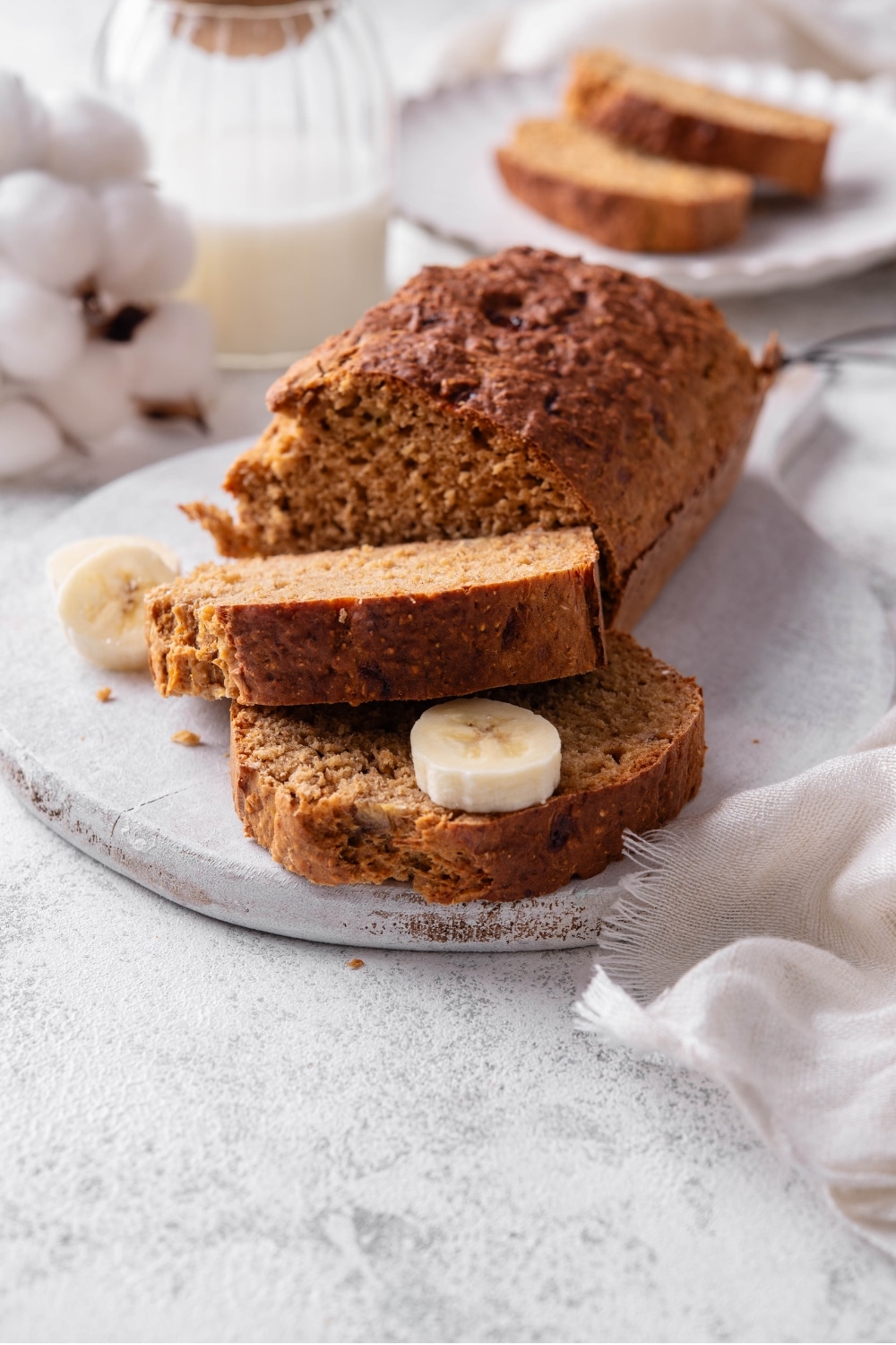 A low calorie banana bread loaf and two slices of banana bread sit on a white serving board. There is a piece of banana in between the two slices, and additional pieces of banana on the board.