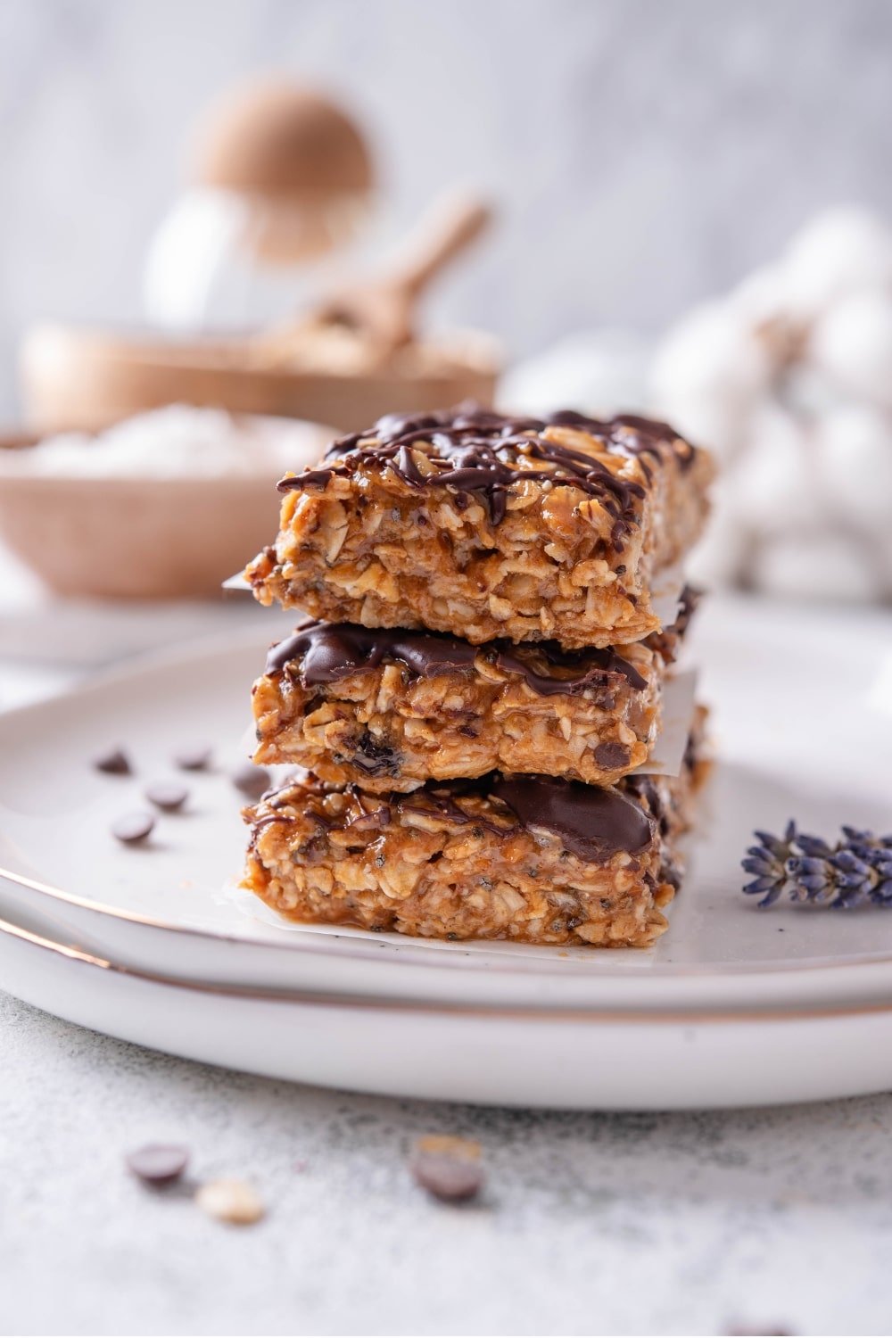 Three low calorie energy bars with chocolate drizzle stacked on a white ceramic plate. There are chocolate chips and oats sprinkled in front of the plate.