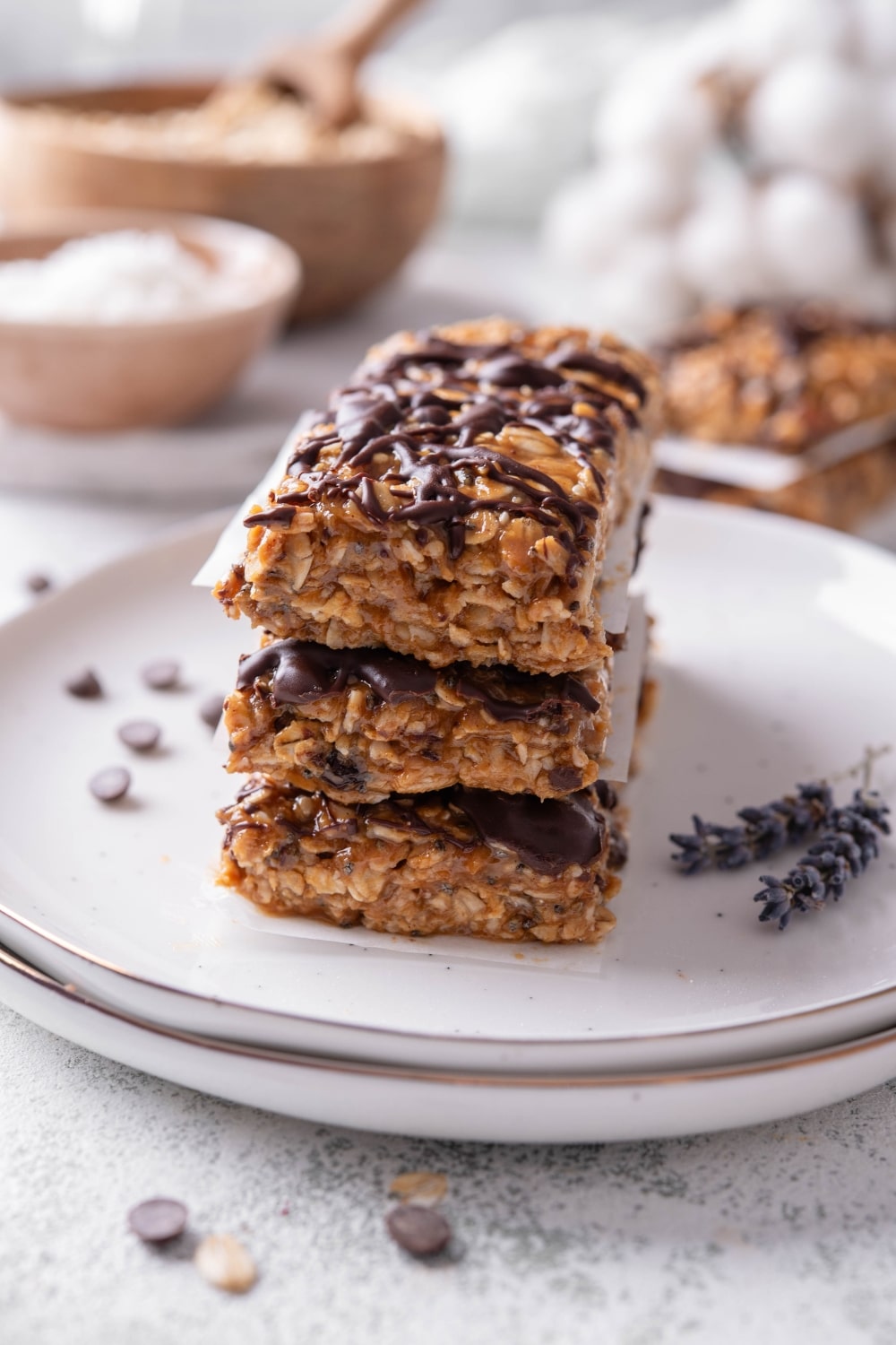 Three low calorie energy bars with chocolate drizzle stacked on a white ceramic plate. There are chocolate chips and oats sprinkled in front of the plate.