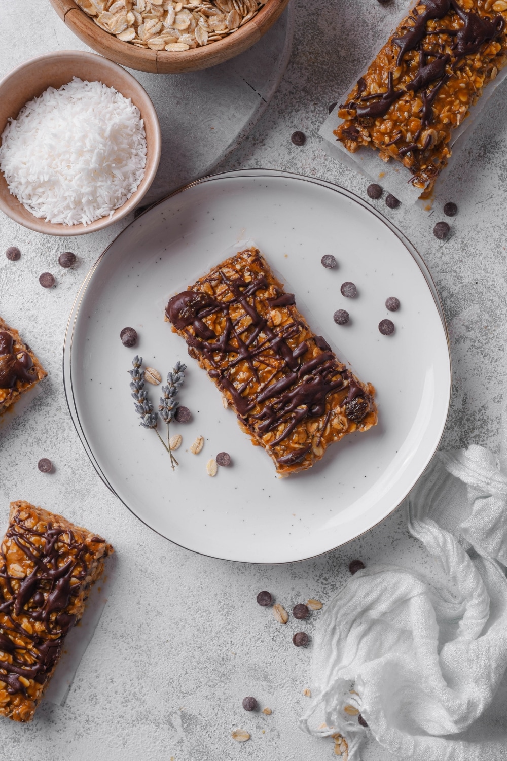 An overhead shot of a low calorie energy bar drizzled with chocolate sauce served on a white plate. There are additional energy bars next to the plate with chocolate chips sprinkled around the plate