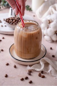 A ribbed glass filled with low calorie iced coffee on a ceramic saucer. A hand is stirring the coffee with a red and white striped straw.
