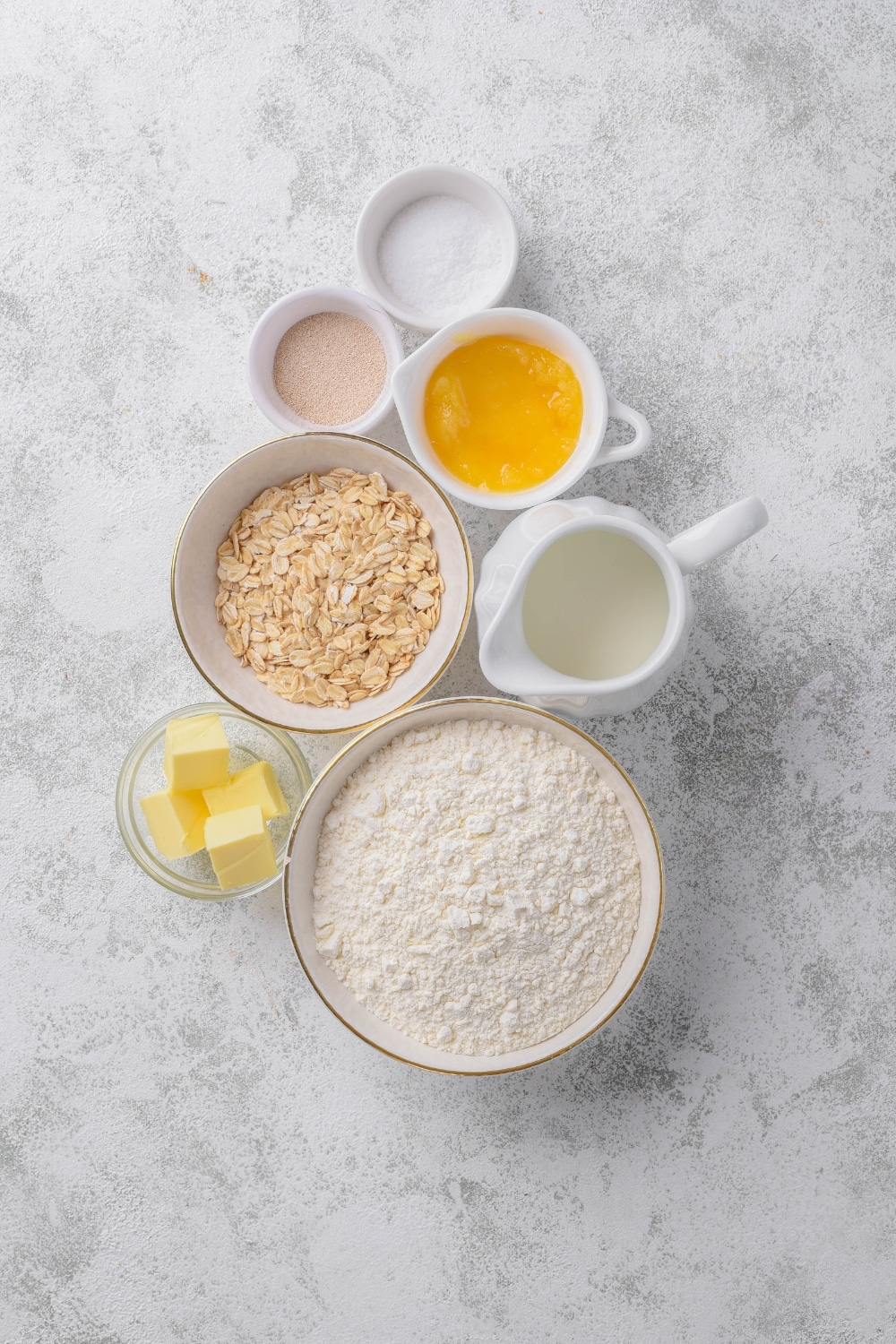 An overhead shot of several bowls of various shapes and sizes holding ingredients for oat bread such as oats, flour, yeast, eggs, milk, and butter.