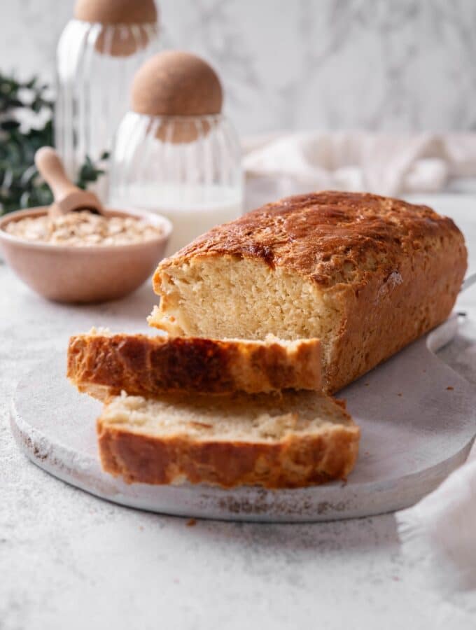 One loaf with two slices of oat bread sit on a wooden server In the background there is a bowl of oats with a scooper inside it.