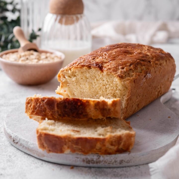 One loaf with two slices of oat bread sit on a wooden server In the background there is a bowl of oats with a scooper inside it.