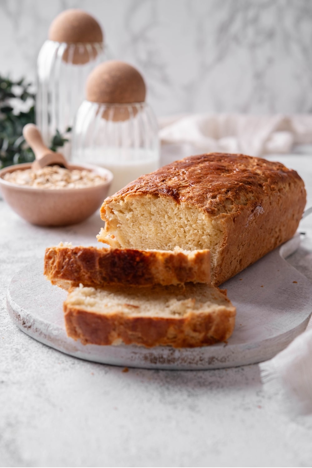 One loaf with two slices of oat bread sit on a wooden server In the background there is a bowl of oats with a scooper inside it.