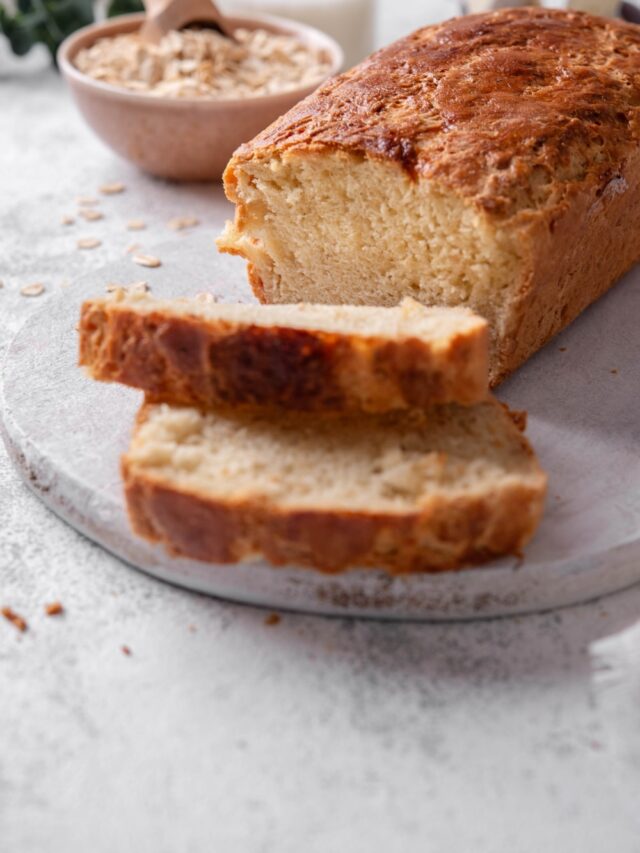 One loaf with two slices of oat bread sit on a wooden server. In the background there is a bowl of oats with a scooper inside it.