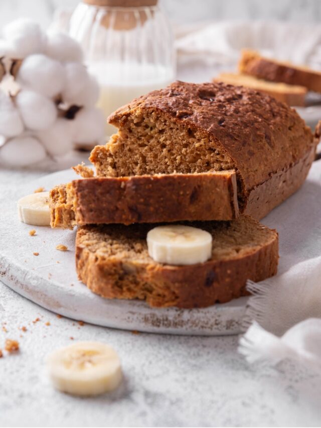 A low calorie banana bread loaf and two slices of banana bread sit on a white serving board. There is a piece of banana in between the two slices, and additional pieces of banana on the board.