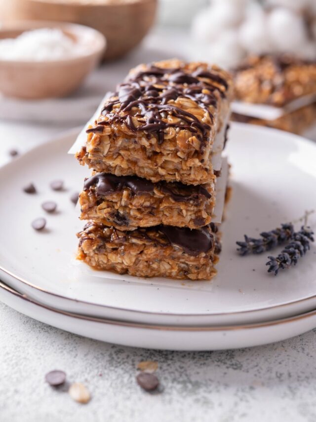 Three low calorie energy bars with chocolate drizzle stacked on a white ceramic plate. There are chocolate chips and oats sprinkled in front of the plate.