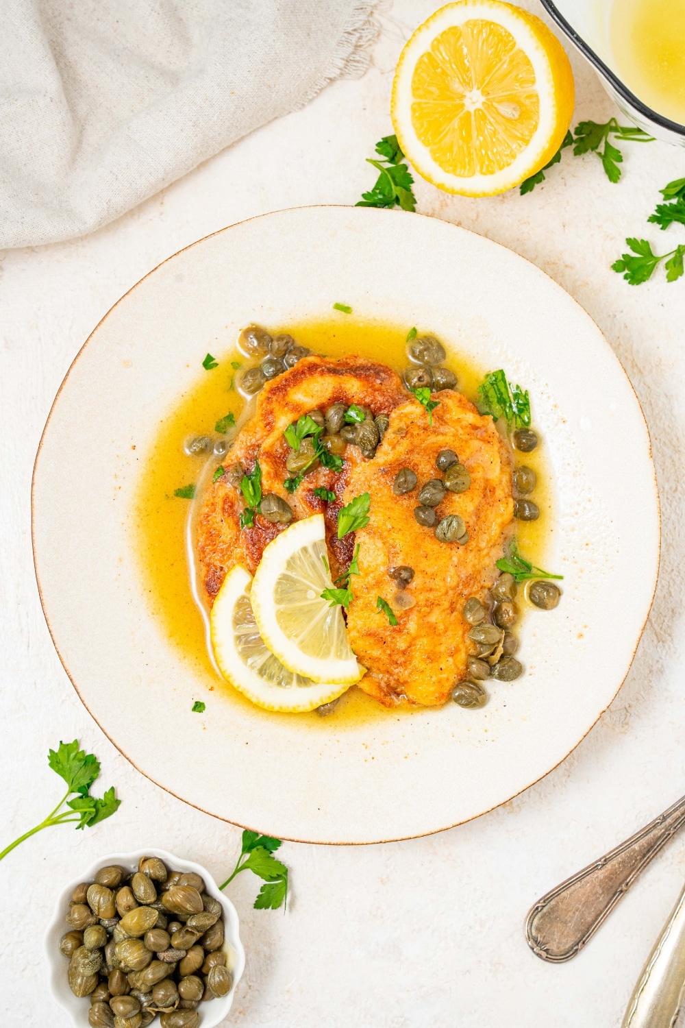 An overhead shot of a plate of two fried calamari steaks with a lemon caper sauce. The plate is garnished with sliced lemon and fresh parsley. There is a bowl of capers next to the plate.