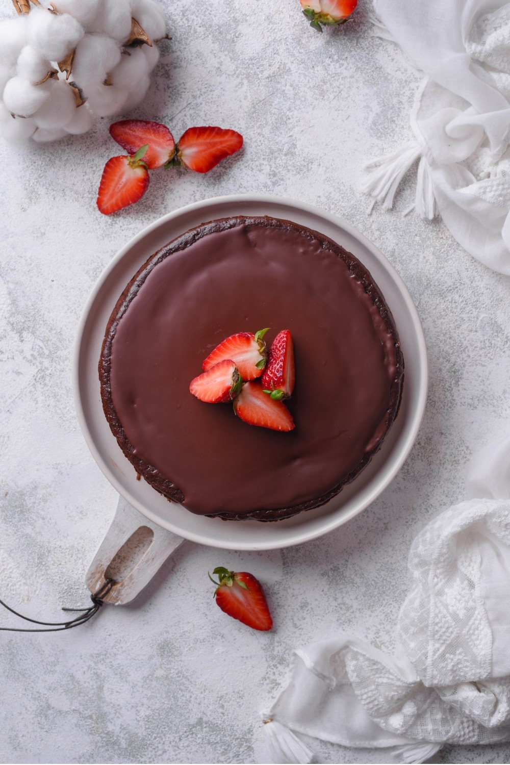 An overhead shot of a whole chocolate protein cake on a white serving board. The cake is topped with sliced strawberries with additional strawberries scattered around it.