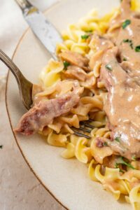 A fork is taking a bite out of crock pot cube steak with egg noodles. There is a small piece of cube steak and some noodles on the fork with a knife in the background.