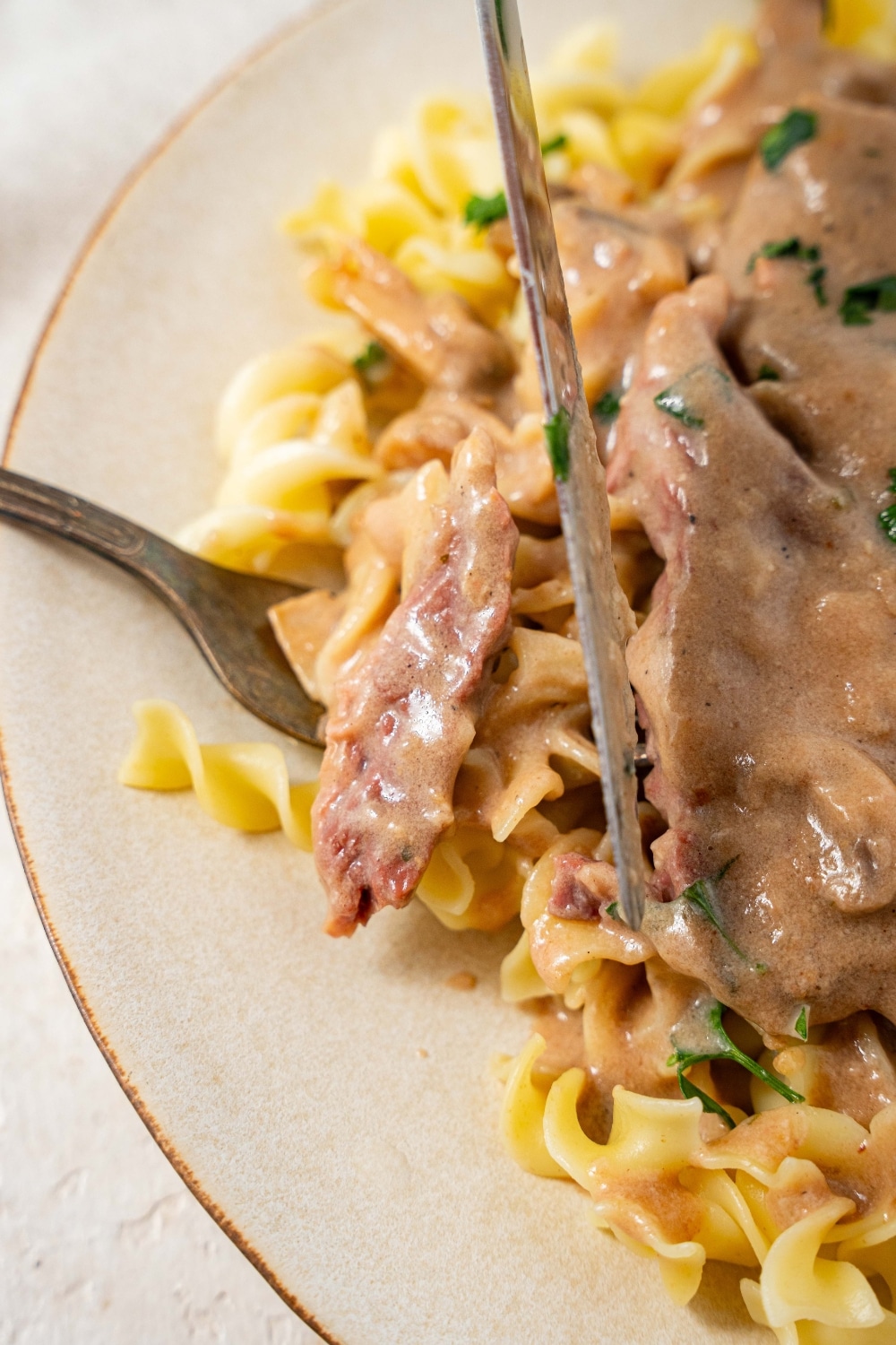 A fork is slicing a bite out of crock pot cube steak with egg noodles. There is a small piece of cube steak and some noodles on the fork with a knife slicing the steak.