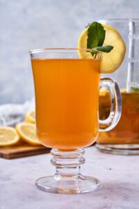 A glass tea mug filled with flu bomb tea garnished with fresh mint and a slice of lemon. There is a glass pitcher of flu bomb tea and a wooden board with sliced lemon in the background.