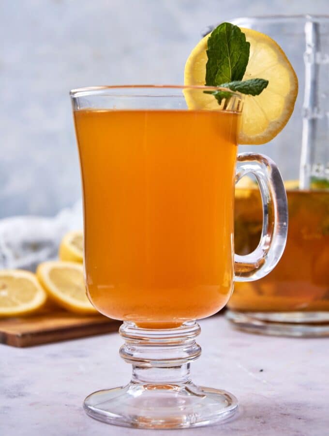 A glass tea mug filled with flu bomb tea garnished with fresh mint and a slice of lemon. There is a glass pitcher of flu bomb tea and a wooden board with sliced lemon in the background.