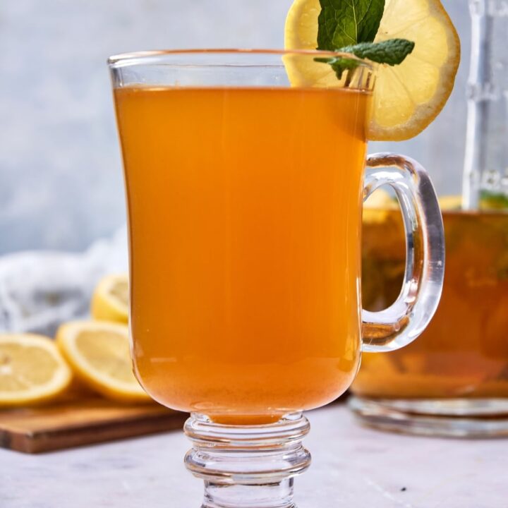 A glass tea mug filled with flu bomb tea garnished with fresh mint and a slice of lemon. There is a glass pitcher of flu bomb tea and a wooden board with sliced lemon in the background.