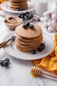 A stack of five high fiber pancakes on a white ceramic plate with a fork to the side. The pancakes are topped with blueberries and there re some blueberries on the plate and off to the side.
