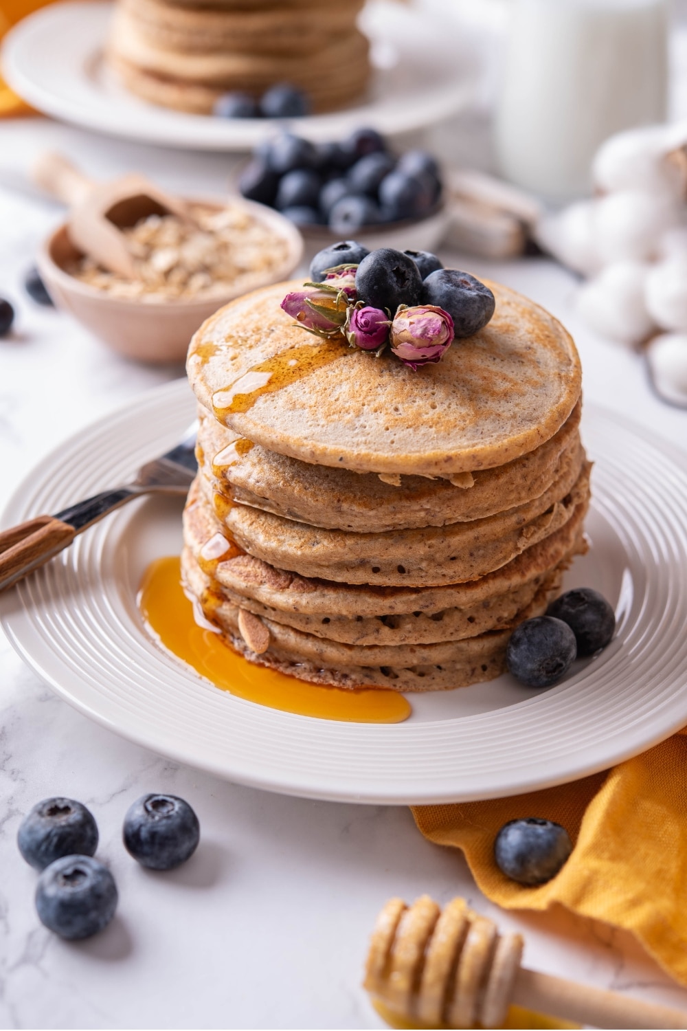 A stack of five high fiber pancakes drizzled with syrup on a white ceramic plate. There is a fork to the side of the stack and the pancakes are topped with blueberries and dried flower buds.