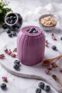 A ribbed glass filled with a high fiber smoothie served on a wooden board. The smoothie is topped with blueberries and chia seeds and there are blueberries and dried flower buds scattered around the board.