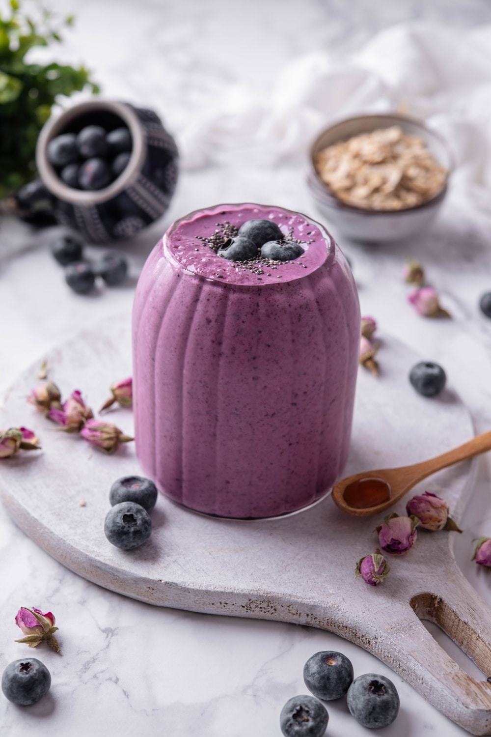 A ribbed glass filled with a high fiber smoothie served on a wooden board. The smoothie is topped with blueberries and chia seeds and there are blueberries and dried flower buds scattered around the board.