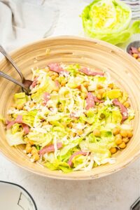 An overhead shot of a large salad bowl filled with the la scala chopped salad. There is iceberg lettuce tossed with salami, chick peas, and shredded mozzarella cheese with a fork and spoon in the salad.