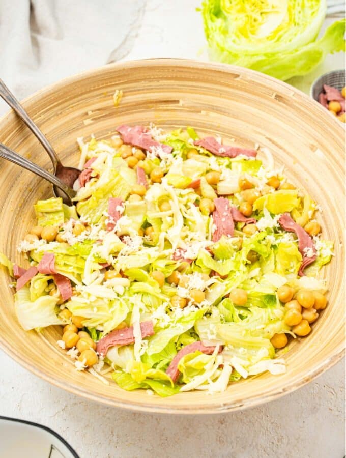 An overhead shot of a large salad bowl filled with the la scala chopped salad. There is iceberg lettuce tossed with salami, chick peas, and shredded mozzarella cheese with a fork and spoon in the salad.