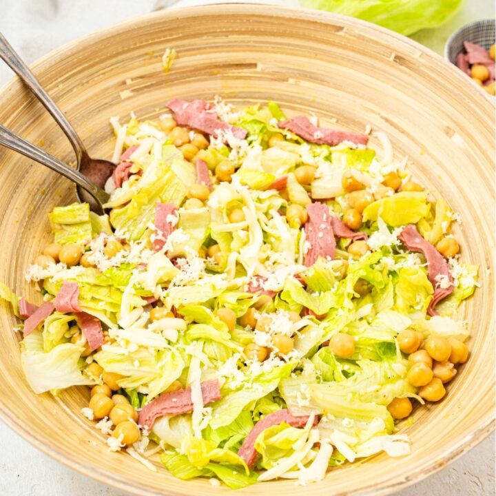 An overhead shot of a large salad bowl filled with the la scala chopped salad. There is iceberg lettuce tossed with salami, chick peas, and shredded mozzarella cheese with a fork and spoon in the salad.