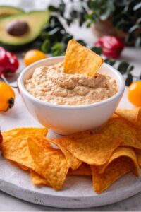 A bowl of low calorie queso dip on a wooden server. There is a tortilla chip inserted into the dip and additional chips around the bowl.