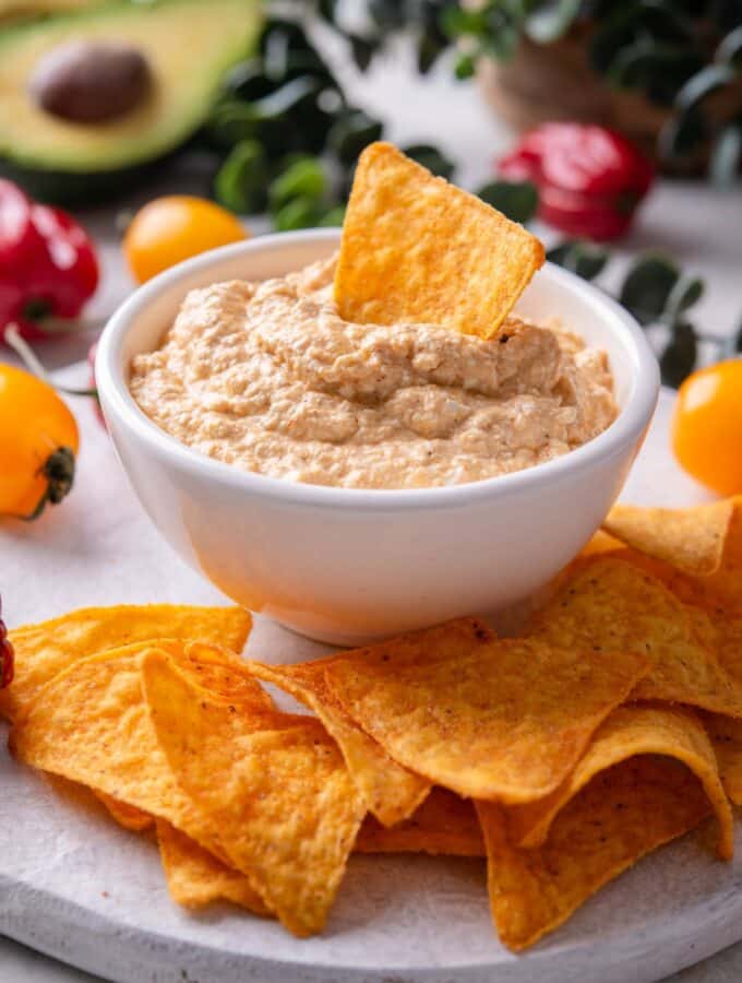 A bowl of low calorie queso dip on a wooden server. There is a tortilla chip inserted into the dip and additional chips around the bowl.