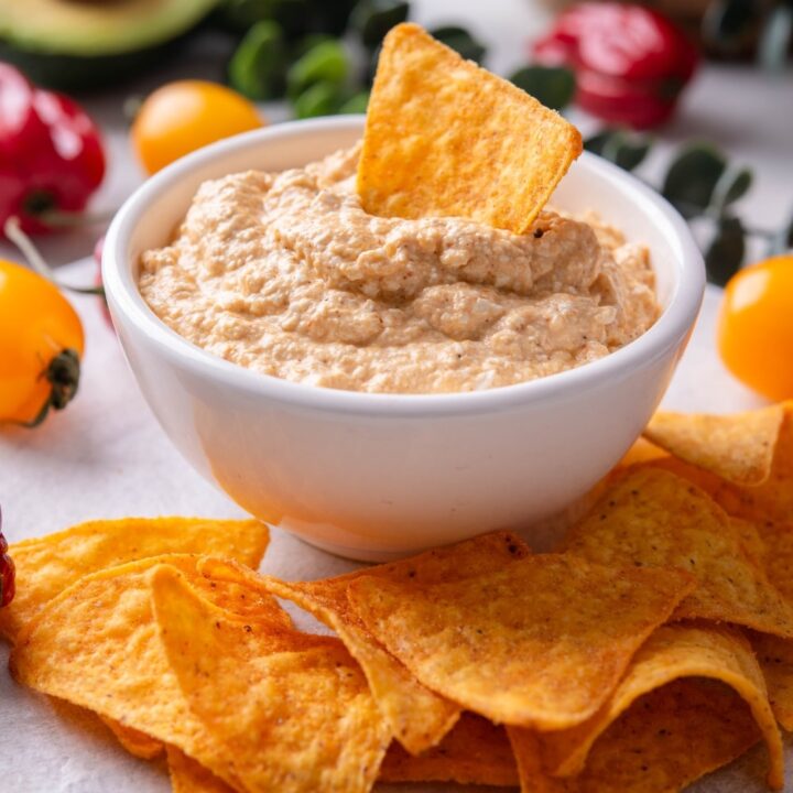 A bowl of low calorie queso dip on a wooden server. There is a tortilla chip inserted into the dip and additional chips around the bowl.