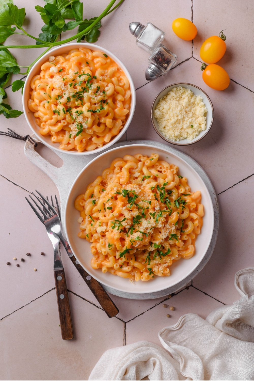 An overhead shot of two bowls of protein mac and cheese. The larger bowl is on a wooden server with two forks next to it. Each bowl is garnished with parsley and grated cheese.