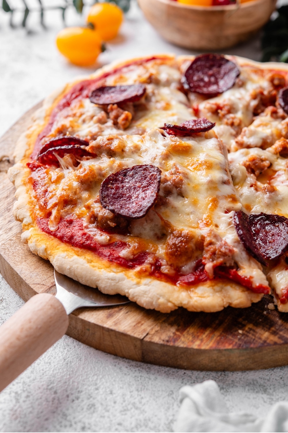 A whole protein pizza topped with cheese, sausage, and pepperoni on a wooden server. There is a pizza server inserted under a slice of pizza.