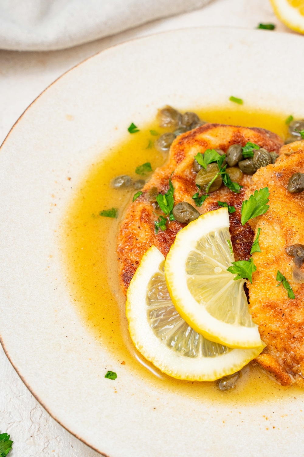 A partial shot of a plate of two fried calamari steaks with a lemon caper sauce. The plate is garnished with sliced lemon and fresh parsley.