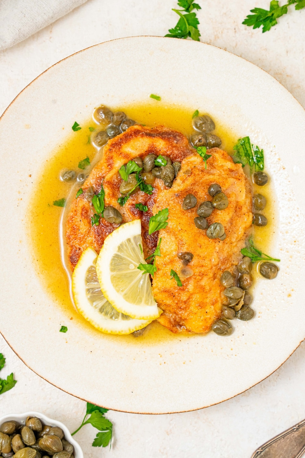 An overhead shot of a plate of two fried calamari steaks with a lemon caper sauce. The plate is garnished with sliced lemon and fresh parsley. There is a bowl of capers next to the plate.