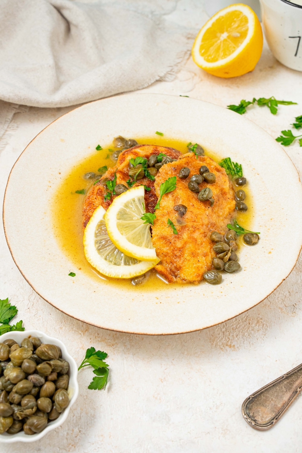 A plate of two fried calamari steaks with a lemon caper sauce. The plate is garnished with sliced lemon and fresh parsley. There is a bowl of capers next to the plate.