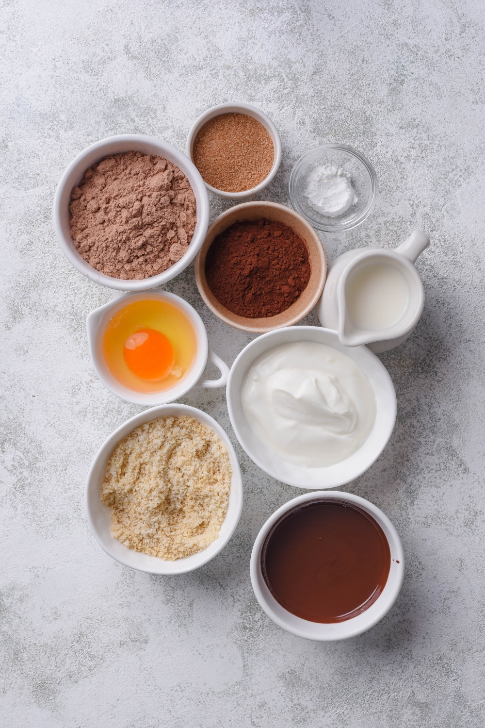 An overhead shot of several bowls containing various ingredients to make a chocolate protein cake including greek yogurt, chocolate protein powder, cocoa powder, almond milk, melted chocolate ganache, almond flour, and a raw egg.