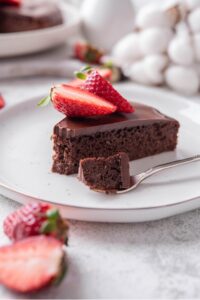 A slice of chocolate protein cake topped with sliced strawberries on a white ceramic plate. There is a fork with a bite of the cake on the plate.