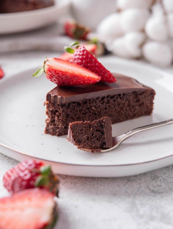A slice of chocolate protein cake topped with sliced strawberries on a white ceramic plate. There is a fork with a bite of the cake on the plate.