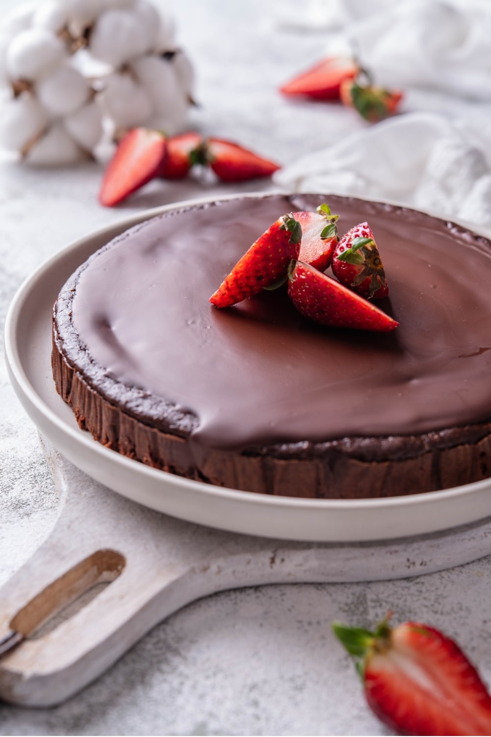 A whole chocolate protein cake topped with sliced strawberries. The cake is served on a white ceramic plate that sits on a white wooden serving board.