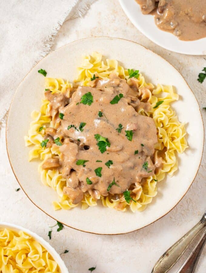 An overhead shot of a plate filled with crock pot cubed steak and gravy on a bed of egg noodles with fresh parsley on top. There is an additional plate of egg noodles and an additional plate of cube steak in gravy on either side.
