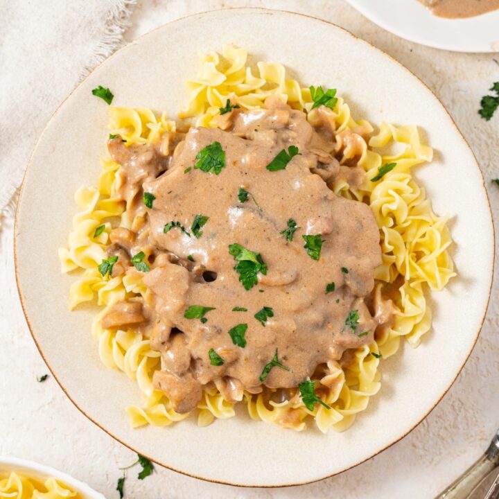 An overhead shot of a plate filled with crock pot cubed steak and gravy on a bed of egg noodles with fresh parsley on top. There is an additional plate of egg noodles and an additional plate of cube steak in gravy on either side.