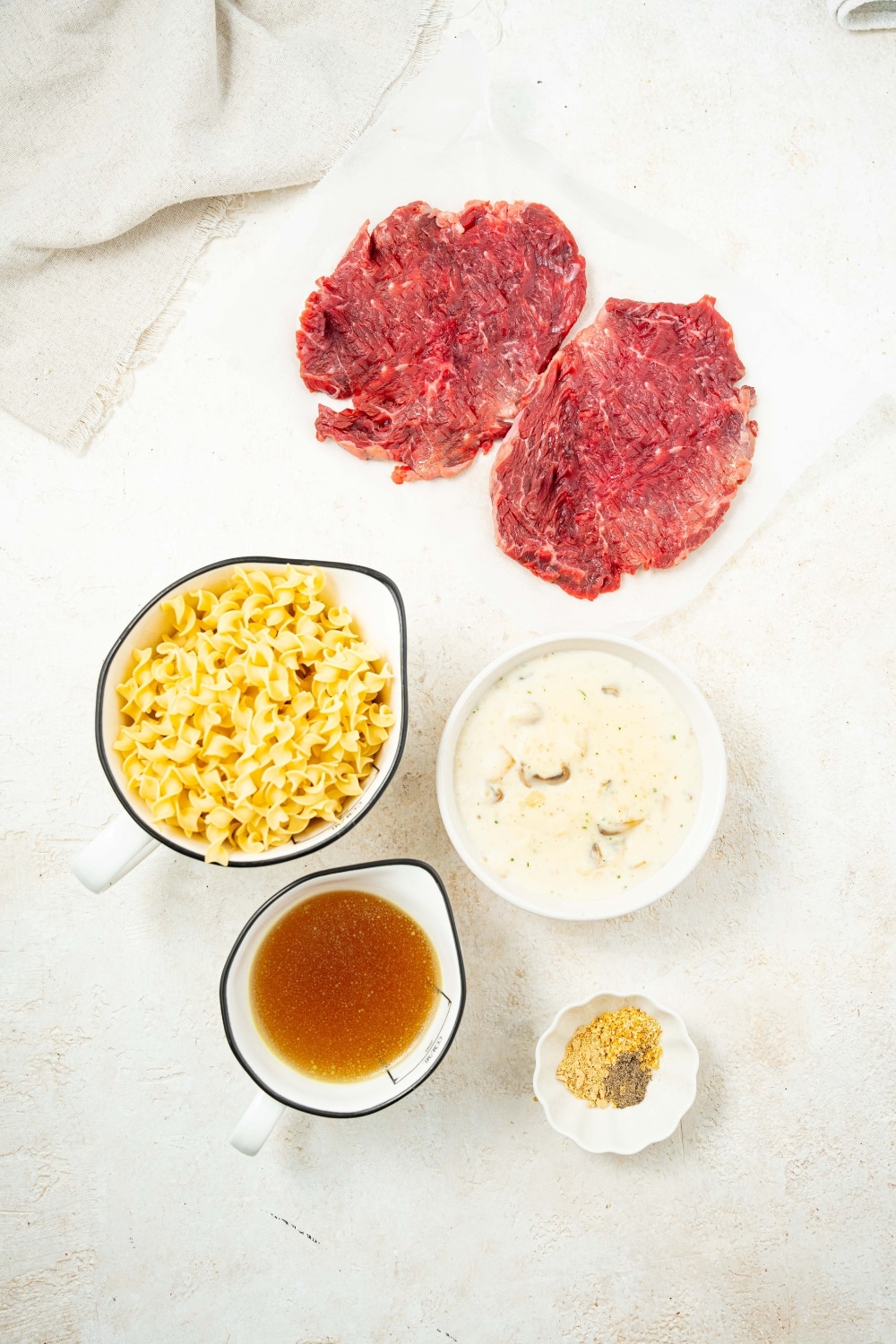 An overhead shot of several ingredients to make crock pot cube steak including a bowl of cream of mushroom soup, a ramekin of seasonings, two cube steaks, and a measuring cup filled with egg noodles and a measuring cup filled with beef broth.